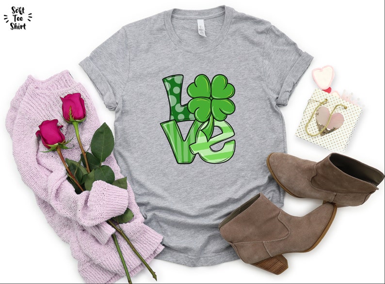 May include: A gray t-shirt with a green "LOVE" graphic featuring a four-leaf clover. The shirt is displayed on a white background with a pair of brown boots, a pink sweater, and a small gift bag.