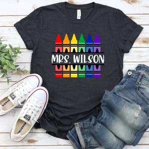 May include: A dark grey t-shirt with a design of colorful crayons and the text "MRS. WILSON" printed on it.