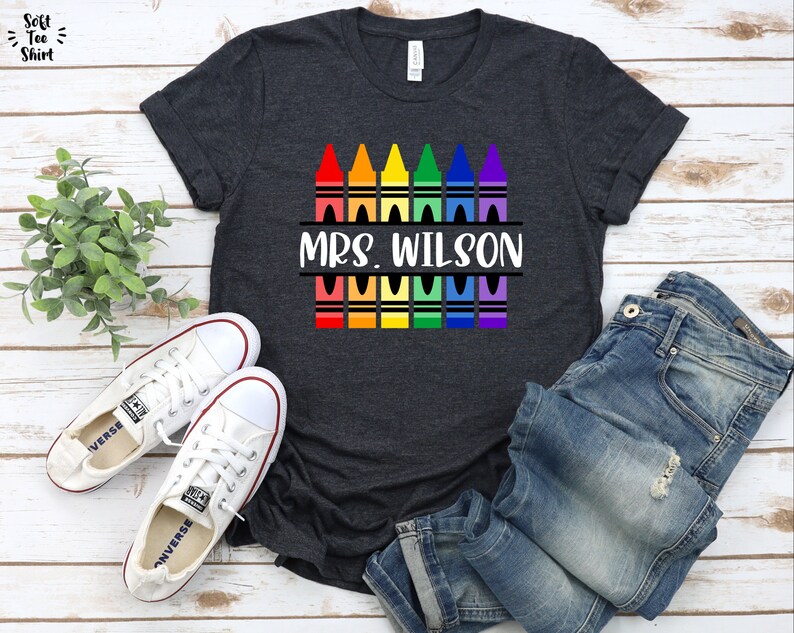 May include: A dark grey t-shirt with a design of colorful crayons and the text "MRS. WILSON" printed on it.