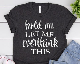 May include: Dark gray t-shirt with rolled-up sleeves. The shirt features the white text "hold on LET ME overthink THIS" in a cursive font. The shirt is paired with blue jeans and white sneakers.