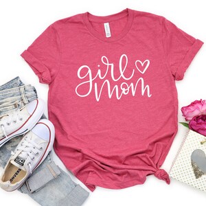 May include: A pink t-shirt with the text "girl mom" in white script with a small heart next to the word "girl".