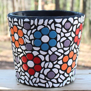 May include: A mosaic-tiled flower pot with a black interior and rim. The exterior features a pattern of white, orange, red, blue, and purple flower designs. The pot is cylindrical and sits on a wooden surface.