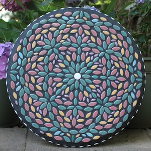 May include: Round mosaic stepping stone with a dark gray background. The mosaic features a floral pattern with colorful leaf-shaped tiles in shades of teal, pink, yellow, and blue. A white border surrounds the edge, and a white circle is in the center.