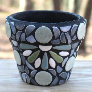 May include: A small, black mosaic flower pot. The pot is decorated with a mosaic pattern of gray, green, and white tiles. The tiles are arranged in a floral design around the pot. The pot is sitting on a wooden surface.