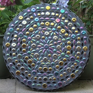 May include: A round, dark gray mosaic stepping stone with a floral design. The mosaic features iridescent glass pieces in gold, silver, blue, and purple. The stepping stone is a decorative garden element.