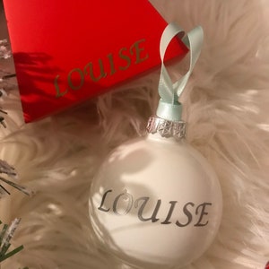 Puede incluir: Adorno navideño blanco con el nombre "Louise" escrito en letras plateadas.