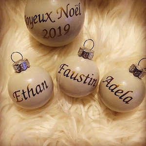 May include: Four white Christmas ornaments with gold lettering. The top ornament says "Joyeux Noel 2019". The other three ornaments say "Ethan", "Faustine", and "Adela".