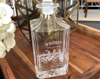 May include: A clear glass decanter with a square base and a stopper. The decanter is etched with the text "Whiskey for my men" and a design of cowboys riding horses.