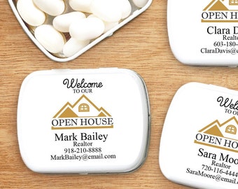 May include: A silver tin filled with white mints, next to three white rectangular tins with gold text that reads "Welcome to our Open House" and realtor contact information. The tins are on a wooden surface.