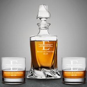May include: A clear glass decanter with a decorative stopper, flanked by two matching lowball glasses. Each piece is engraved with the name "EMMANUEL", "Dexter & Catherine" and the date "05.29". The decanter and glasses are filled with amber liquid.
