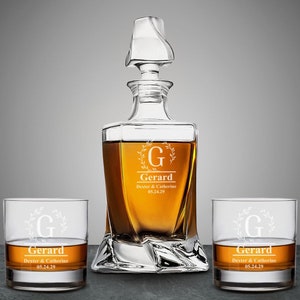 May include: A clear glass decanter with a stopper and two matching glasses. The decanter and glasses are engraved with the letter "G" and the name "Gerard" along with the date "05.24.29".