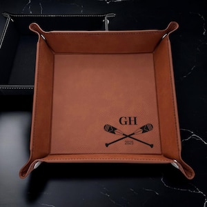 May include: A brown leather valet tray with a personalized inscription "GH 2025" and a pair of crossed oars.