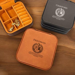 May include: Three square jewelry cases in brown, black, and orange leather. The open brown case reveals an orange interior with ring slots and compartments. The black case has white stitching and the name "Crawford" printed on it. The orange case has the name "Friedman" printed on it.