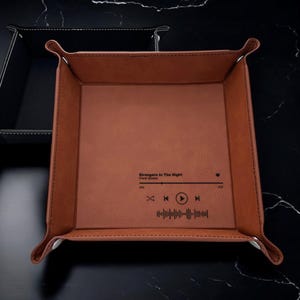 May include: A brown leather catchall tray with a black music player graphic and the text "Strangers in The Night" by Frank Sinatra. The tray has a square shape and is made of a soft, supple leather.