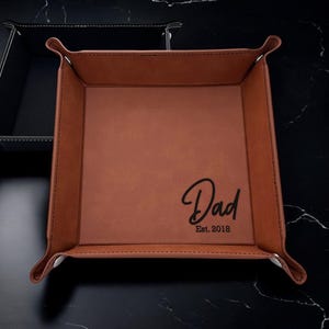 May include: A brown leather valet tray with a black inscription that reads "Dad Est. 2018".
