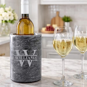 May include: A cylindrical gray marble wine chiller engraved with the name WILLIAMS and a W monogram, accompanied by two clear glass wine glasses, each featuring the same white engraved WILLIAMS name and W monogram design.