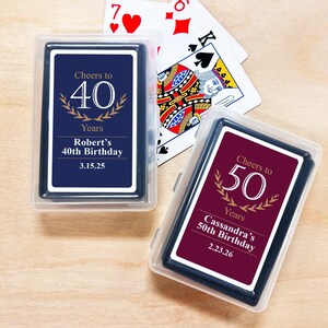 May include: Two decks of playing cards in clear plastic cases. One deck is navy blue with "Cheers to 40 Years" and the name "Robert" and a date. The other deck is burgundy with "Cheers to 50 Years" and the name "Cassandra" and a date. Playing cards are scattered around.
