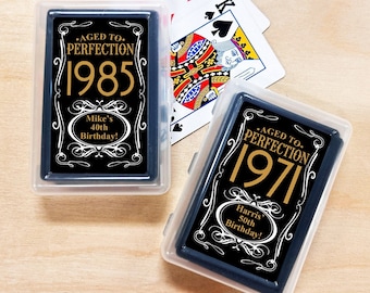 May include: Two clear plastic boxes containing decks of playing cards. The cards have a black background with gold lettering. The text on the cards reads "Aged to Perfection" and the year 1985 or 1971. The text also includes the name "Mike's" or "Harris'" and the phrase "40th Birthday!" or "50th Birthday!"