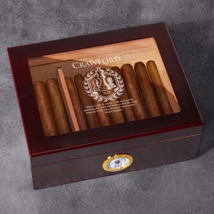 May include: A dark brown wooden cigar box with a glass top. The box has a gold-colored metal hinge and a gold-colored metal clock face on the front. The box is engraved with the name "Crawford" and a chess piece design. The quote "Class holds master in its own hands, shackling the world and brain so over the near freedom of the very strongest must suffer." - Albert Einstein is also engraved on the box.