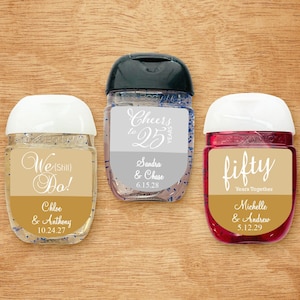 May include: Three hand sanitizer bottles with personalized labels. The labels are gold and white with different anniversary messages. The first bottle says "We (Still) Do!" with the names Chloe & Anthony and the date 10.24.27. The second bottle says "Cheers to 25 Years" with the names Sandra & Chase and the date 6.15.28. The third bottle says "fifty Years Together" with the names Michelle & Andrew and the date 5.12.29.