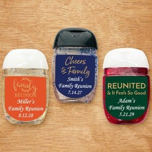 May include: Three clear plastic hand sanitizer bottles with colorful labels. The labels have gold text and say "family reunion" with the date of the reunion. The bottles are on a wood background.