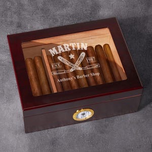 May include: A dark wood cigar humidor with a glass top, containing several cigars. The humidor has a brass-colored hygrometer and the text "MARTIN EST. 1987 Anthony's Barber Shop" is visible on the inside.