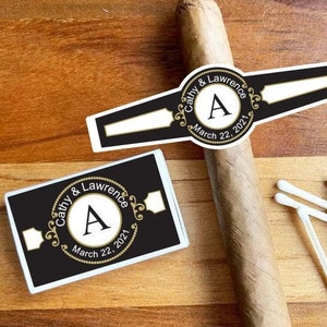 May include: A black and gold cigar band with the text "Cathy & Lawrence" and the letter "A" in a circle. The date "March 22, 2021" is also included. A matchbox with the same design is also shown.