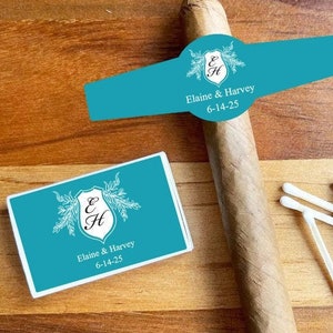 May include: Teal blue wedding favor box with a white border and a white design of a shield with the letters E and H inside. The box has the text "Elaine & Harvey 6-14-25" printed on it. A teal blue cigar band with the same design and text is also shown.
