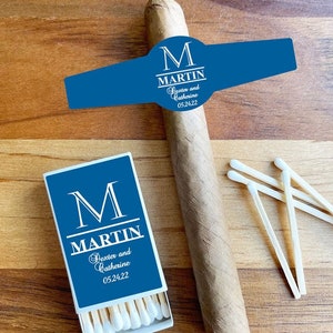 May include: A blue and white personalized cigar band with the text "M MARTIN Dexter and Catherine 05.24.22". A blue and white matchbox with the same text. The matchbox contains white matches.