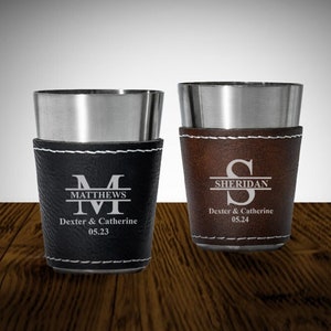 May include: Two stainless steel shot glasses with leather sleeves. The left glass has a black sleeve with the text "MATTHEWS" and "Dexter & Catherine 05.23". The right glass has a brown sleeve with the text "SHERIDAN" and "Dexter & Catherine 05.24".
