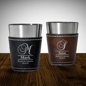 May include: Two stainless steel shot glasses with leather wraps. The left glass has a black leather wrap with the name "Mark" and the initial "M" engraved. The right glass has a brown leather wrap with the name "Sean" and the initial "S" engraved.