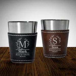 May include: Two stainless steel cups with leather wraps. The cups are engraved with a monogram, name, and date. The left cup has a black leather wrap and the right cup has a brown leather wrap.