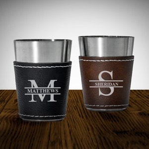 May include: Two stainless steel shot glasses with leather wraps. The left glass has a black leather wrap with the letter "M" and the name "MATTHEWS" engraved on it. The right glass has a brown leather wrap with the letter "S" and the name "SHERIDAN" engraved on it.