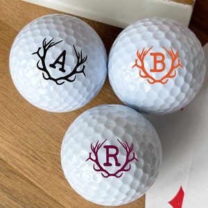 May include: Three white golf balls with black, orange, and purple antlers and letters A, B, and R in the center.