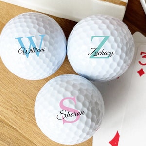 May include: Three white golf balls with personalized names and initials. The names are William, Zachary, and Sharon. The initials are W, Z, and S. The initials are in different colors: blue, teal, and pink.