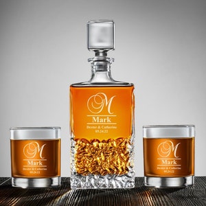 May include: A personalized crystal decanter and two matching glasses. The decanter and glasses are engraved with the letter "M", the name "Mark", and the text "Dexter & Catherine 05.24.22".