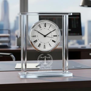 May include: A silver-framed desk clock with a white clock face and black Roman numerals. The clock has a silver base and a clear glass frame. The clock face has the name "Richard" and a scales of justice symbol.