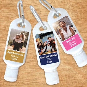 May include: Three white plastic bottles with carabiner clips. Each bottle has a personalized label with a photo and text. The first bottle says "Happy Anniversary Gilmores 5.12.28". The second bottle says "Happy 40th Birthday Paul 2.2.27". The third bottle says "Cheers to 50 Georgina 11.23.26".