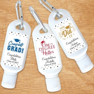 May include: Three white plastic bottles with black and white labels. The labels have graduation-themed text and graphics. The bottles have a carabiner clip attached to the top. The text on the labels reads "Congrats Grad! Congratulations Blake Class of 2027", "One Degree Hotter Congratulations Michelle Class of 2028", and "You Did It Congratulations Andrew Class of 2029".