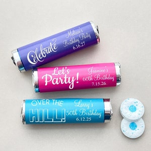 May include: Three personalized mini chocolate bars wrapped in foil with different birthday messages. The bars are purple, pink, and blue. The text on the purple bar reads "Celebrate Melissa's Birthday Party 6.16.27". The text on the pink bar reads "Let's Party! Jasmine's 60th Birthday 7.15.26". The text on the blue bar reads "Over the Hill! Larry's 50th Birthday 6.12.25".