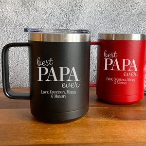 May include: Two stainless steel travel mugs with black and red powder coated finishes. Both mugs have a white inscription that reads "best PAPA ever" with the names "Courtney, Miles & Mommy" underneath.