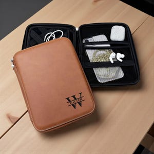 May include: A brown leather tech organizer with a zipper closure, open to reveal a black interior with a phone, earbuds, and other accessories. The organizer has the name "WALTER" printed on the front.