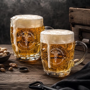 May include: Two clear glass beer mugs filled with golden beer and thick white foam. Each mug features an etched caduceus symbol, the text "DR. JAMES FRIEDMAN" and "SAVING LIVES SINCE 2008" in a circular design on the front surface of the glass.