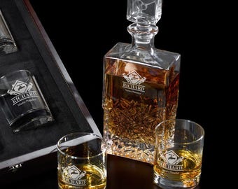 May include: A clear glass decanter with a square stopper and two matching lowball glasses, all filled with amber liquid. The decanter and glasses are etched with the name "RICHARD" and a logo. The set is displayed on a black surface.