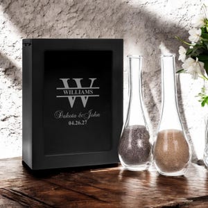May include: Black wooden box with a glass lid for a sand ceremony. The box is engraved with the text "W WILLIAMS Dakota & John 04.26.27". Two clear glass bottles with sand inside are in front of the box.