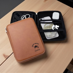 May include: A brown leather tech organizer with a zipper closure, open to reveal a black interior with a phone, earbuds, and other accessories. The organizer has the text "BEST SKIER JASPER" printed on the front.