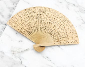 May include: A light brown wooden hand fan with intricate cutouts and a white tassel. The fan is closed and lying on a white marble surface.