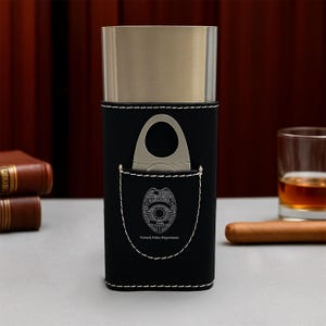 May include: A black leather cigar case with a silver-colored metal top and white stitching. A cigar cutter is in the front pocket. The case has the Newark Police Department badge engraved on it. A cigar and a glass of amber liquid are in the background.