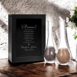 May include: Black wooden box with a glass panel displaying a wedding vow inscription. The inscription reads "Promise" and includes the names Rachel & Joshua and the date May 12, 2028. Two clear glass sand ceremony vases with gray and tan sand are in front of the box.