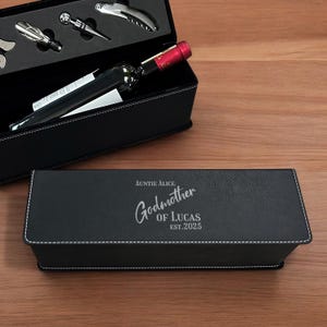 May include: A black wine gift set with a bottle of wine and wine tools. The set includes a black box with the words "Auntie Alice Godmother of Lucas Est. 2025" in silver lettering. The box is on a wooden surface.